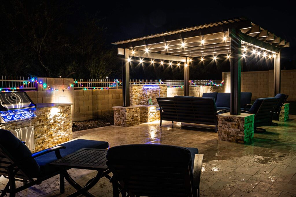 A resort style backyard at night with a waterfall, beautiful lighting, a pergola, and a firepit. - ProNail Outdoor Living