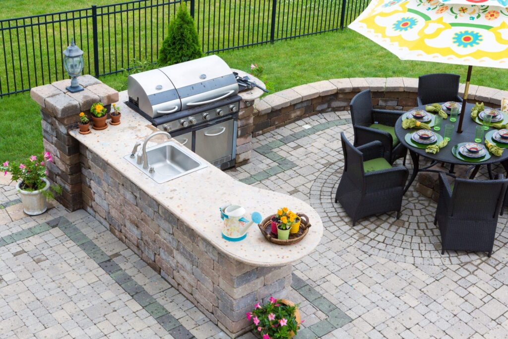 High angle view of a stylish outdoor kitchen, gas barbecue and dining table set for entertaining guests with formal place settings and flowers on a paved patio (High angle view of a stylish outdoor kitchen, gas barbecue and dining table set for entertaining.) - Outdoor Living